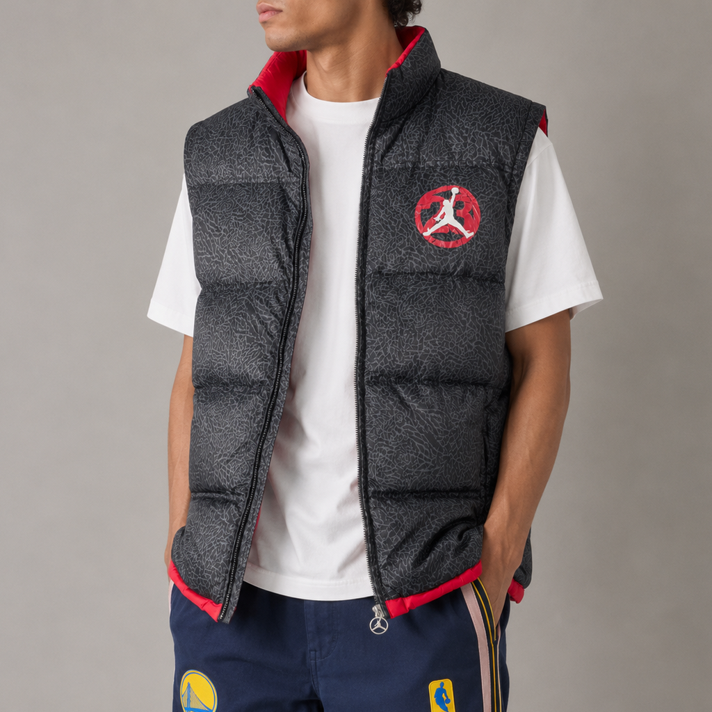 A model wearing a dark gray textured puffer vest with red lining and a red Jordan logo on the chest, layered over a white t-shirt and navy blue track pants.